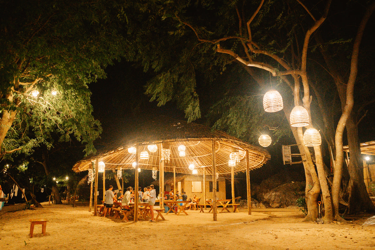 Guests at dinner under a bamboo pavilion hung with woven lanterns at a Keelooma beach camp at night, Palawan